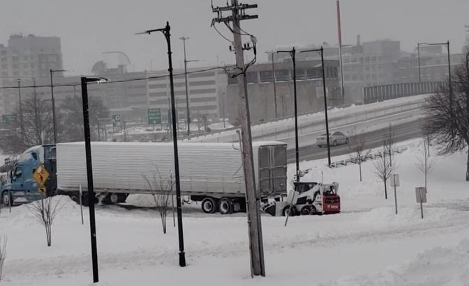 Small loader helps free large semi-truck stuck in snow by pushing truck from behind in Boston